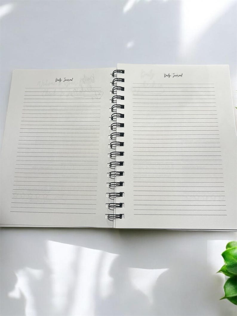 May include: An open spiral bound notebook with lined pages. The notebook is white with black text that says "Daily Journal" on both sides.