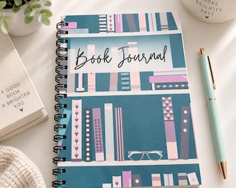 May include: A spiral-bound book journal with a teal cover featuring a graphic illustration of bookshelves filled with various books in shades of purple, pink, and blue. The text Book Journal is printed in black script on the front cover.
