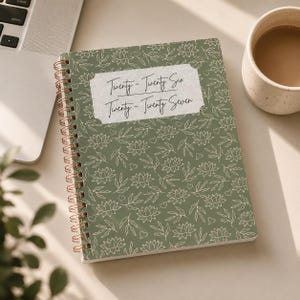 May include: A spiral-bound notebook with a sage green cover featuring a white line-art pattern of lotus flowers, leaves, and small hearts. A central white label displays the text Twenty-Twenty Six and Twenty-Twenty Seven in black script.