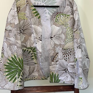 May include: A floral print kimono-style jacket with a V-neck. The jacket features a botanical pattern in shades of gray, white, and green. The jacket is hanging on a black hanger.