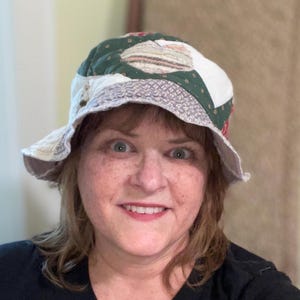 May include: A patchwork bucket hat with a wide brim. The hat features a mix of fabrics in green, white, and patterned designs. The brim is a light beige color. The hat is worn by a person.