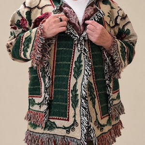 May include: A long-sleeved tapestry jacket featuring a woven pattern of dark green leaves and deep red berries on a beige background. The garment has a collared neckline, front pockets, and decorative fringe trim along the edges and cuffs.