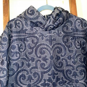 Pu&ograve; includere: Giacca con cappuccio blu navy con un motivo floreale ricamato bianco. La giacca &egrave; appesa a una gruccia blu su uno sfondo di scala in legno. Il design della giacca presenta un motivo a riccioli e foglie ripetuto.