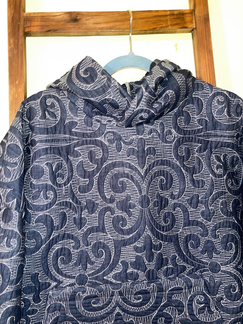 Pu&ograve; includere: Giacca con cappuccio blu navy con un motivo floreale ricamato bianco. La giacca &egrave; appesa a una gruccia blu su uno sfondo di scala in legno. Il design della giacca presenta un motivo a riccioli e foglie ripetuto.