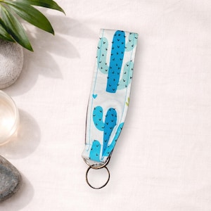 Könnte beinhalten: Ein weißes Schlüsselband aus Stoff mit einem sich wiederholenden Muster aus blauen Kakteen. Das Schlüsselband hat einen Metallring und ist etwa 15 cm lang. Das Design umfasst hellblaue und dunkelblaue Kakteen mit kleinen Herzakzenten.
