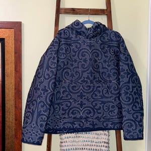 Pu&ograve; includere: Giacca trapuntata blu navy con un motivo floreale bianco ripetuto. La giacca ha un collo alto ed &egrave; appesa a un gancio blu. La giacca &egrave; esposta su una scala di legno.