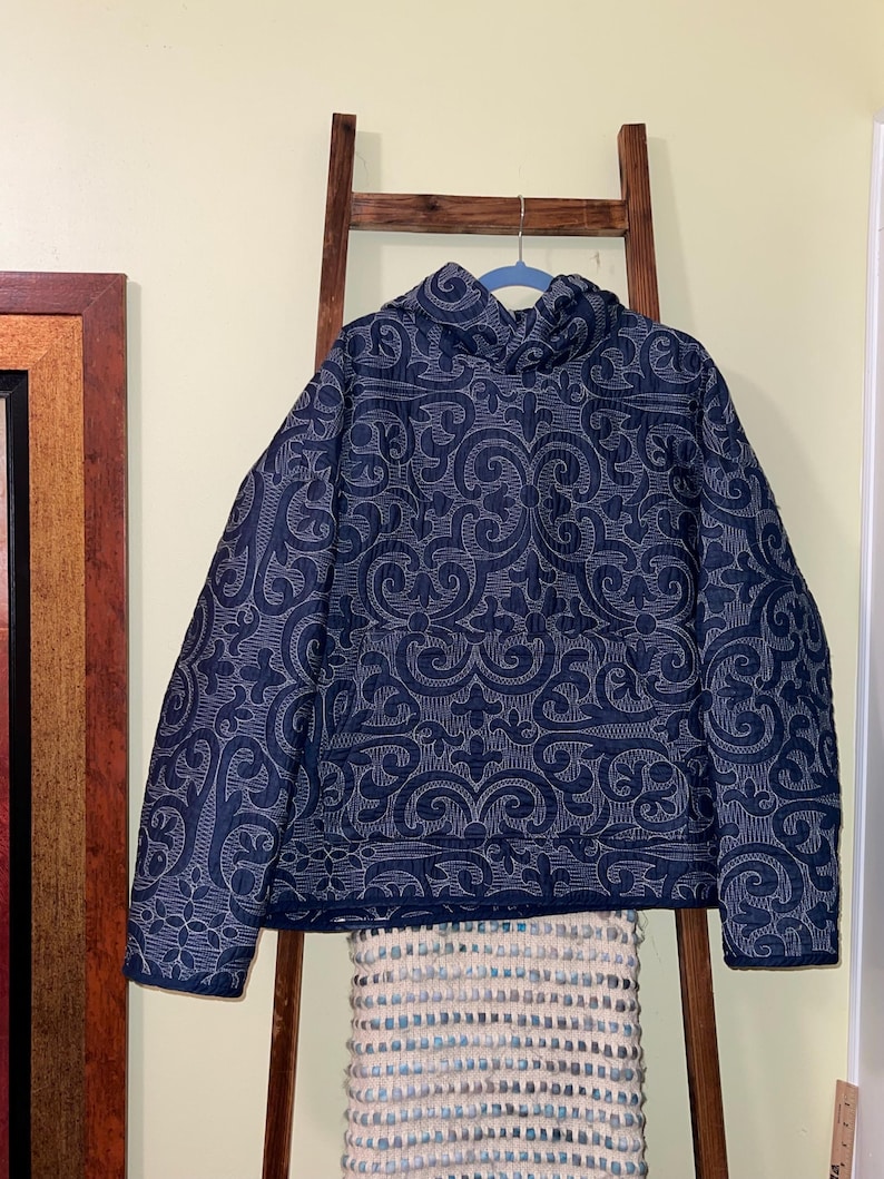 Pu&ograve; includere: Giacca trapuntata blu navy con un motivo floreale bianco ripetuto. La giacca ha un collo alto ed &egrave; appesa a un gancio blu. La giacca &egrave; esposta su una scala di legno.