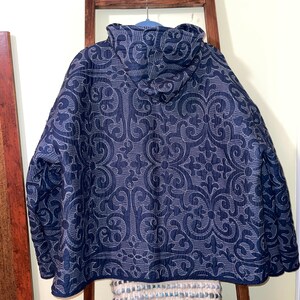 Pu&ograve; includere: Giacca trapuntata blu navy con cappuccio, con un intricato motivo a volute bianche. La giacca &egrave; appesa a un appendiabiti blu. Il design della giacca &egrave; visibile da dietro. La giacca &egrave; di colore blu scuro.