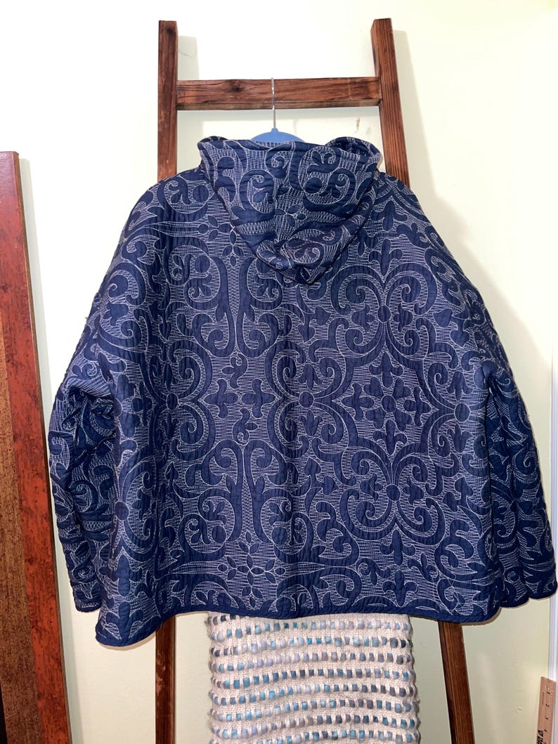 Pu&ograve; includere: Giacca trapuntata blu navy con cappuccio, con un intricato motivo a volute bianche. La giacca &egrave; appesa a un appendiabiti blu. Il design della giacca &egrave; visibile da dietro. La giacca &egrave; di colore blu scuro.