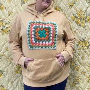 May include: Mustard-colored hoodie with a large, square, crocheted patch on the front. The patch features a colorful granny square design with shades of green, pink, orange, and white. The hoodie has a kangaroo pocket and a drawstring hood.