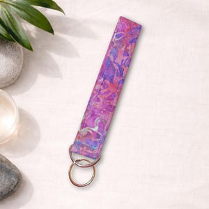 May include: A vibrant, patterned wristlet key fob in shades of pink, purple, and blue. The fabric features a watercolor-like design with a silver key ring attached. The wrist strap is approximately 6 inches long.