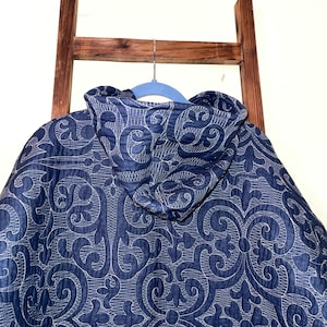 Pu&ograve; includere: Giacca trapuntata blu navy con cappuccio, con un motivo bianco e ornato. La giacca &egrave; appesa a una gruccia blu, con una scala di legno sullo sfondo.