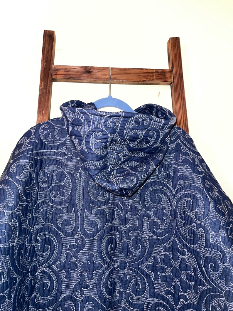 Pu&ograve; includere: Giacca trapuntata blu navy con cappuccio, con un motivo bianco e ornato. La giacca &egrave; appesa a una gruccia blu, con una scala di legno sullo sfondo.