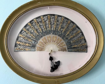 May include: A black and gold lace fan with a white center is displayed in an oval gold frame with a pink mat.
