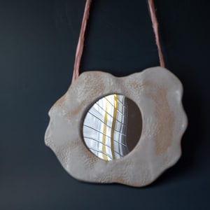 May include: A decorative, off-white ceramic mirror with an irregular, organic shape. The mirror has a round reflective surface and is suspended by a pink cord. The frame has a textured surface with light brown accents.