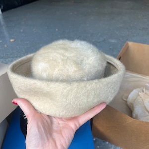 May include: A vintage, cream-coloured hat with a rounded crown and a wide brim. The hat is made of a soft, fuzzy material. The hat is held in a person's hand.