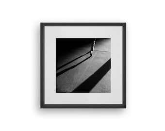 May include: Black and white photograph of a person walking away from the camera, casting a long shadow on the pavement.