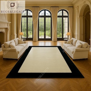 Könnte beinhalten: Ein großer rechteckiger Teppich mit schwarzem Rand und cremefarbenem Mittelteil. Der Teppich befindet sich in einem Wohnzimmer mit zwei cremefarbenen Sofas, großen Fenstern und Parkettböden. Das Logo "RUGS & LEGACY" befindet sich oben links.