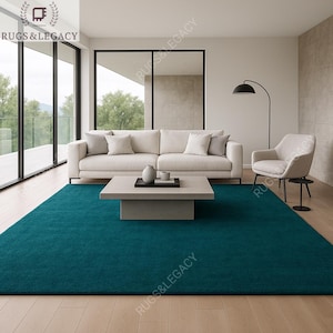May include: A teal-colored area rug dominates the foreground of a modern living room. A white sofa with matching pillows sits against a wall, with a square coffee table in front. The room features large windows and a black floor lamp. The logo "RUGS & LEGACY" is visible.
