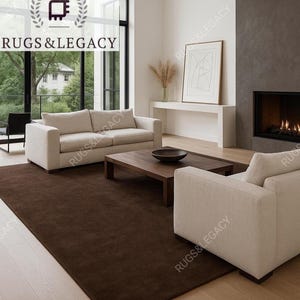 May include: A modern living room featuring a large, dark brown rug. The room includes a beige sofa, a matching armchair, and a wooden coffee table. A fireplace and large windows with a view of trees complete the scene. The brand name "Rugs & Legacy" is visible.