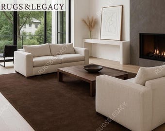 May include: A modern living room featuring a large, dark brown rug. The room includes a beige sofa, a matching armchair, and a wooden coffee table. A fireplace and large windows with a view of trees complete the scene. The brand name "Rugs & Legacy" is visible.