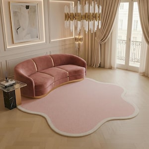 May include: A curved, dusty rose velvet sofa with gold metal accents, paired with a large, irregular, cloud-shaped light pink area rug featuring a cream-coloured border, positioned on a light-toned wooden floor in a bright, neutral-coloured room.