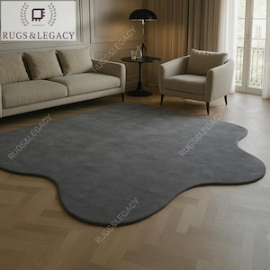 Könnte beinhalten: Ein großer, unregelmäßig geformter grauer Teppich dominiert die Raummitte. Der Teppich hat einen organischen, fließenden Rand. Der Raum hat eine neutrale Farbpalette mit Sofa, Sessel und Holzboden. Der Text "RUGS & LEGACY" ist sichtbar.