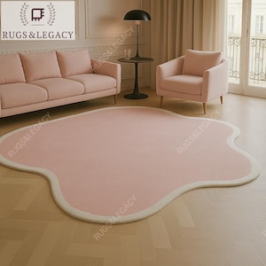Op de afbeelding: Een groot, roze tapijt met een onregelmatige vorm en een crèmekleurige rand. Het tapijt ligt op een lichtgekleurde houten vloer en is omgeven door roze meubels. Het logo "RUGS & LEGACY" staat in de linkerbovenhoek.