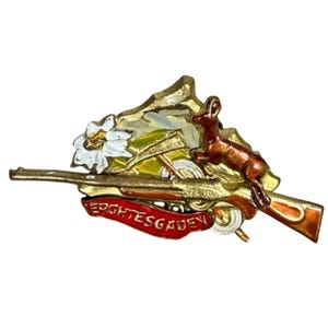 May include: A vintage gold-toned metal pin featuring a rifle, a leaping deer, and a mountain scene with a white flower. The pin has a red banner with the text "EIGHTESGADEN".