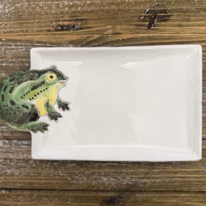 May include: A rectangular white ceramic tray featuring a three-dimensional green frog figure attached to the left side. The frog has yellow accents on its chest and around its eyes, with dark green spots across its back and limbs, contrasting with the smooth, glossy white surface of the tray.