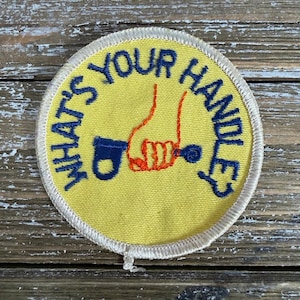May include: Round yellow patch with the text "WHAT'S YOUR HANDLE?" in blue. The patch features a hand holding a blue object with a key. The hand and key details are in orange and blue.