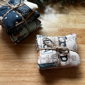 May include: A stack of four small, square fabric sachets tied with twine. The sachets feature various patterns, including a navy blue with white dots, a yellow and white striped design, and a map print.