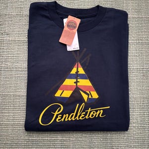May include: Navy blue t-shirt featuring a colorful teepee design with yellow, red, and blue stripes. The word "Pendleton" is written in yellow script below the teepee. A price tag is attached to the shirt.