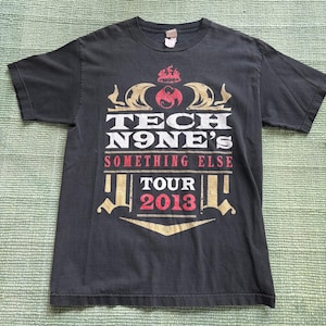 May include: Black t-shirt featuring the text "TECH N9NE'S SOMETHING ELSE TOUR 2013" in white, gold, and red. The design includes a stylized logo and decorative elements. The shirt is made of a soft material.