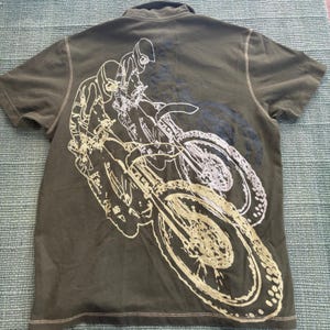 May include: Olive green polo shirt featuring a white and gold graphic of a motocross racer on a motorcycle. The short-sleeved shirt has a collared neckline and a stitched hemline.