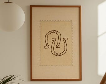 Puede incluir: Una obra de arte enmarcada con un fondo beige y un diseño de herradura negra. La obra de arte está en un marco marrón y cuelga de una pared blanca en un pasillo. Se ve una escalera con escalones de madera y una barandilla blanca.