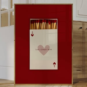 Pode incluir: Obra de arte emoldurada com fundo vermelho e design de carta branca. O cartão exibe um coração rosa, o texto "Love always wins" e o símbolo do Ás de Copas. A parte superior da moldura mostra uma caixa de fósforos.