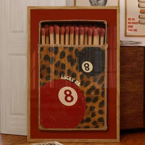 May include: A framed artwork featuring a large matchbox with a leopard print design and the words "Lucky Me" and the number 8. The matchsticks are visible inside the box. Two other framed artworks are visible in the background.