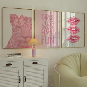 May include: Three framed wall art prints with gold frames. The prints feature a pink leopard with a bow, the text "GIRLS JUST WANNA HAVE FUN", and three pink lip prints. A white cabinet, lamp, and chair are in the foreground.