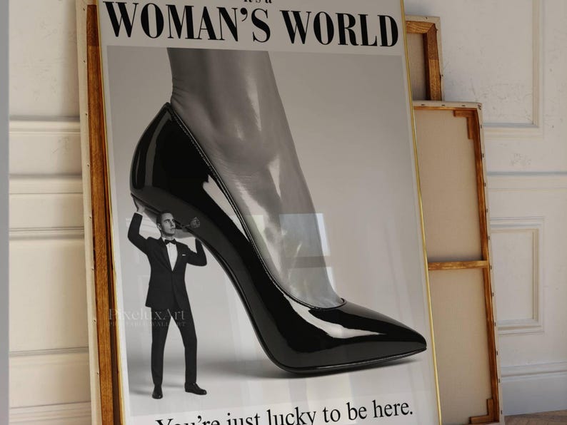 May include: A framed poster with the text "WOMAN'S WORLD" at the top. The image features a giant black high-heeled shoe with a man in a tuxedo holding it up. The text "You're just lucky to be here" is at the bottom.