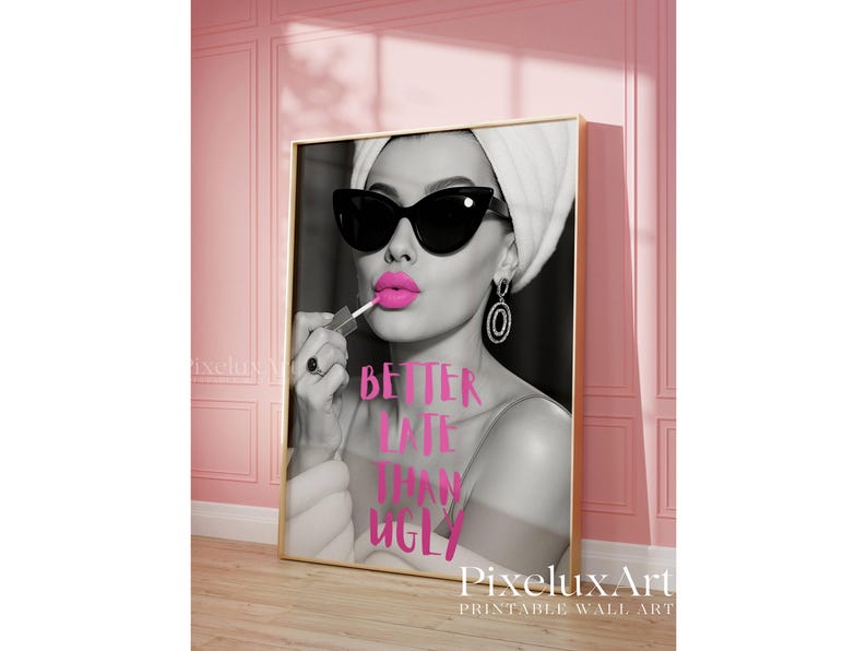 May include: Black and white wall art print featuring a woman applying bright pink lipstick, wearing sunglasses and a towel. The text "BETTER LATE THAN UGLY" is in pink. The print is in a gold frame, leaning against a pink wall.