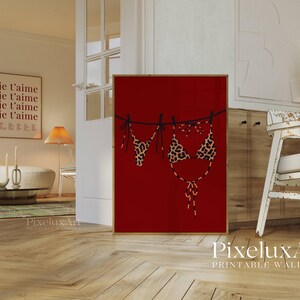 May include: A framed red print featuring a leopard print bikini hanging on a washing line. The artwork is set in a living room with a sofa, table, and other decorative items. The print is in a wooden frame.