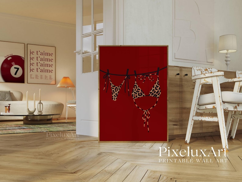 May include: A framed red print featuring a leopard print bikini hanging on a washing line. The artwork is set in a living room with a sofa, table, and other decorative items. The print is in a wooden frame.