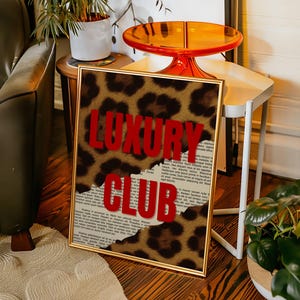 May include: A framed art print with a gold border. The print features a leopard print design with the words "LUXURY CLUB" in bold red letters. The background is a torn newspaper. The artwork is displayed in a room with a green sofa and a small side table.