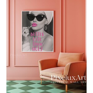 May include: A framed black and white print featuring a woman applying lipstick, wearing sunglasses and a head wrap. The pink text on the print reads "BETTER LATE THAN UGLY". The print hangs on a pink wall above a peach armchair.