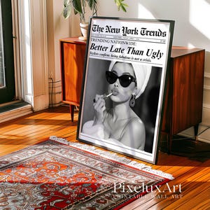 May include: Black and white framed print featuring a woman applying lipstick, wearing sunglasses and a towel on her head. The print is styled as a newspaper front page with the headline "Better Late Than Ugly."