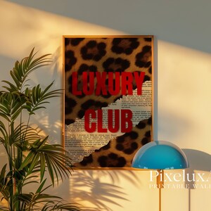 May include: A framed wall art print with a leopard print background and the words "LUXURY CLUB" in bold red letters. The artwork has a torn paper effect, revealing a newspaper-style print underneath. The print is displayed on a wall.