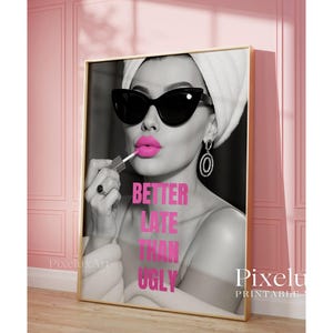 May include: A framed print featuring a black and white image of a woman applying bright pink lipstick. She wears sunglasses, a towel on her head, and large earrings. The text "BETTER LATE THAN UGLY" is in pink. The print is in a gold frame.