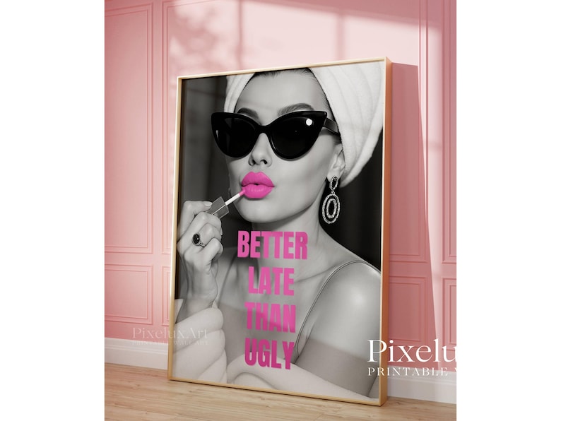 May include: A framed print featuring a black and white image of a woman applying bright pink lipstick. She wears sunglasses, a towel on her head, and large earrings. The text "BETTER LATE THAN UGLY" is in pink. The print is in a gold frame.