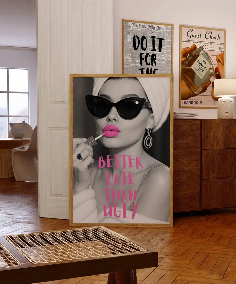 May include: A framed black and white print featuring a woman applying bright pink lipstick, wearing sunglasses and a towel. The text "BETTER LATE THAN UGLY" is written in pink. Other framed prints are visible in the background.